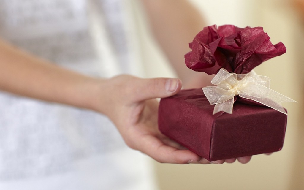 An outstretched hand giving a pretty gift - a small box wrapped in red tissue with a cream ribbon, symbolic of the gift of Health Assessments for Christmas